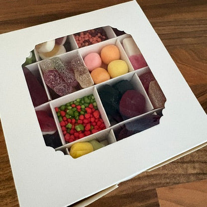 A white cardboard gift box with a window displaying assorted colorful candies and sweets arranged in compartments, photographed on a wooden surface.