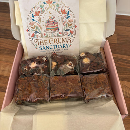 An open pink gift box containing several individually wrapped brownie bars and a printed card for 'The Crumb Sanctuary'.