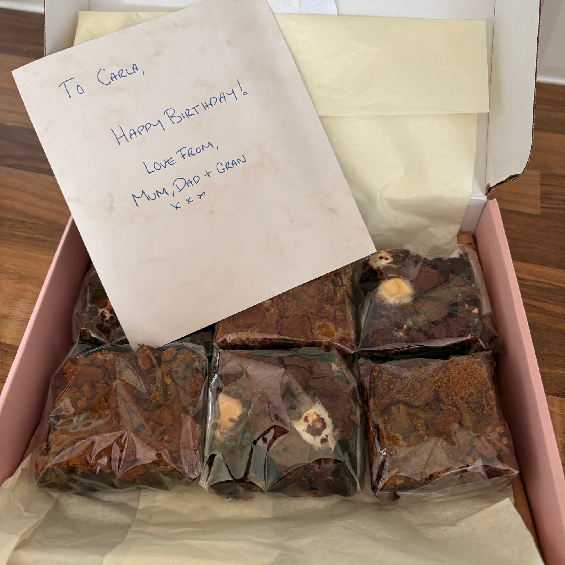 A pink gift box lined with cream tissue paper, containing several individually wrapped brown chocolate treats or cookies. A handwritten note is placed on top of the box.
