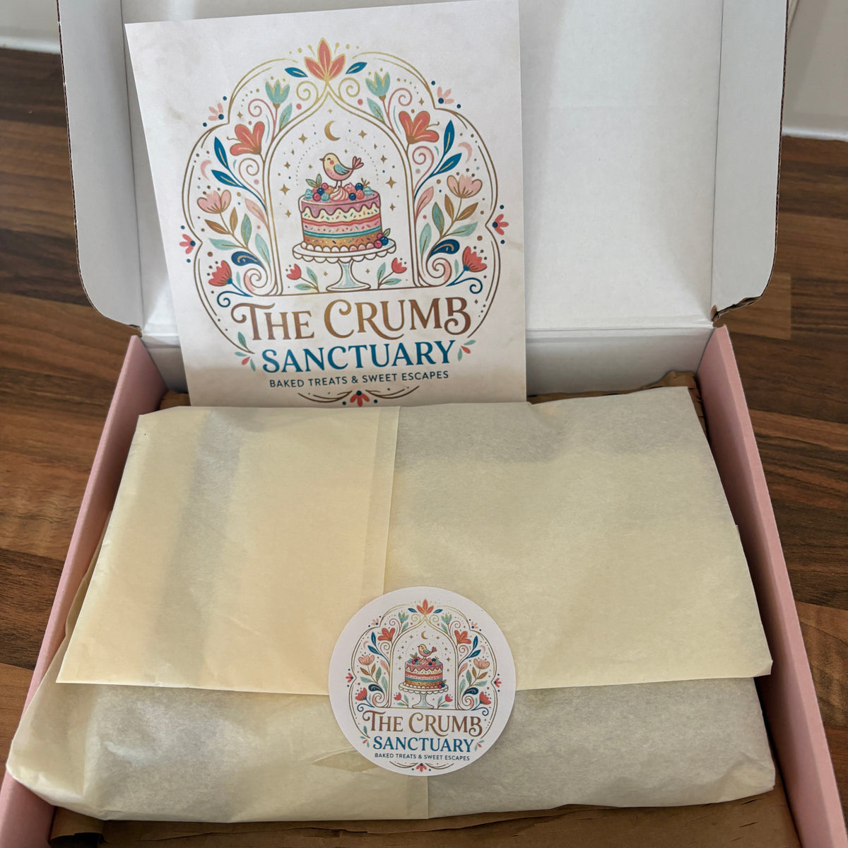 An open pink gift box on a wooden table containing a cream-colored tissue-wrapped item and a printed card with the 'The Crumb Sanctuary' logo. A matching round sticker is visible on the wrap.