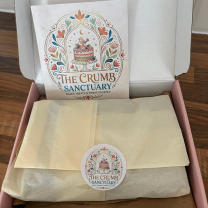 An open pink gift box on a wooden table containing a cream-colored tissue-wrapped item and a printed card with the 'The Crumb Sanctuary' logo. A matching round sticker is visible on the wrap.