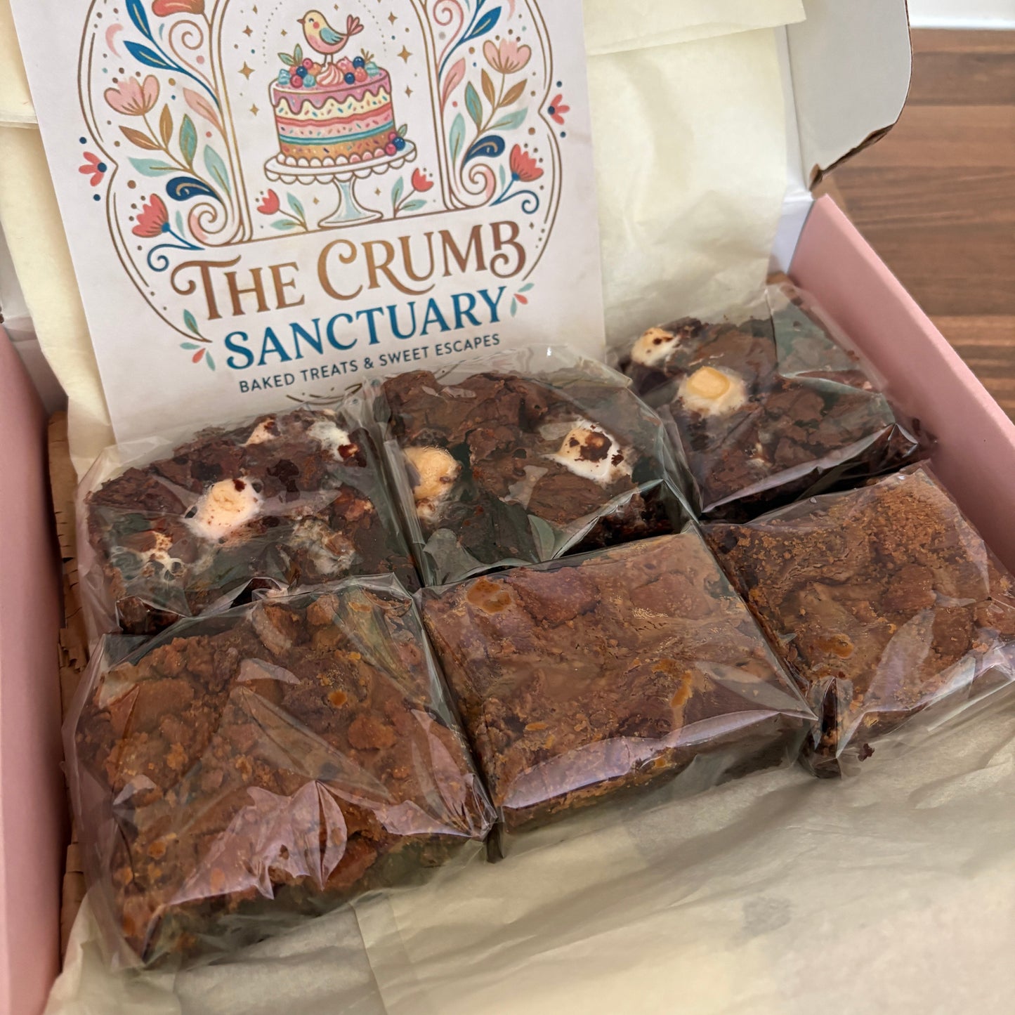 A boxed assortment of chocolate brownies from The Crumb Sanctuary, shown inside a pink box with tissue paper and a branded card.