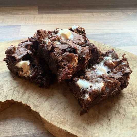 Two rich, dark chocolate rocky road brownies with marshmallow and chocolate digestive chunks, stacked on a rustic wooden cutting board.